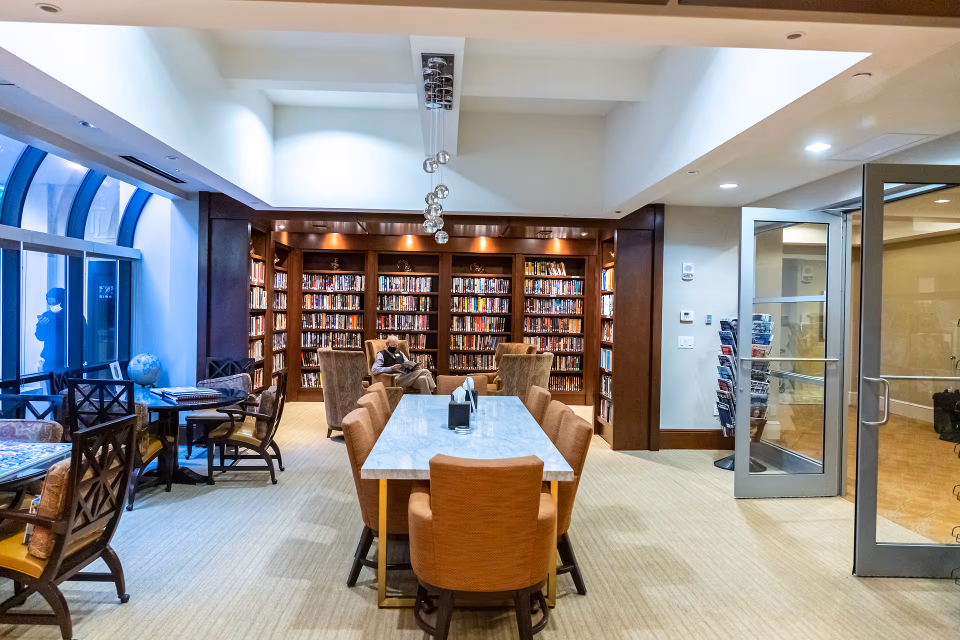 A well-lit library room with wooden bookshelves filled with books along the back wall. In the center, there is a long table with several orange cushioned chairs around it. To the left, there are smaller tables with chairs near large arched windows. A person is seated in a chair reading a book. On the right side, there is a glass door leading to another room.