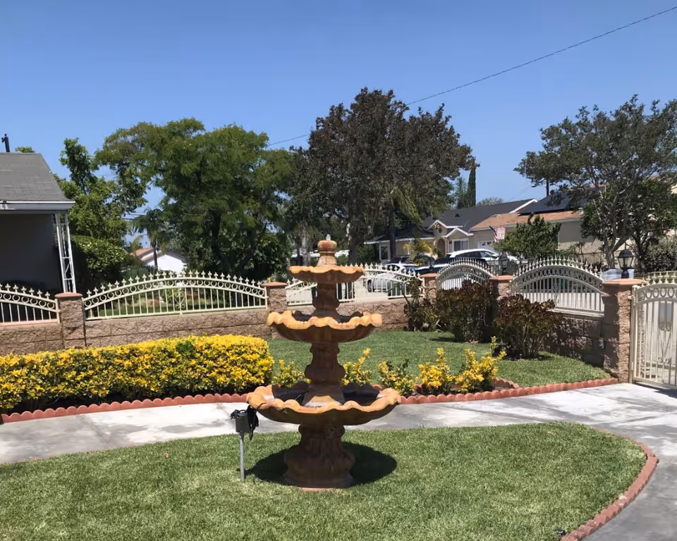 Three-tier stone fountain on a grassy front yard with a decorative fence and neighboring houses in the background.