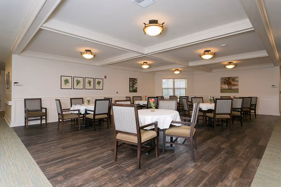 A dining room with several round tables covered with white tablecloths, each surrounded by wooden chairs with cushioned seats and backs. The room has wooden flooring, white walls adorned with framed botanical and landscape artwork, and ceiling lights providing warm illumination. A window with blinds is visible on the far wall.