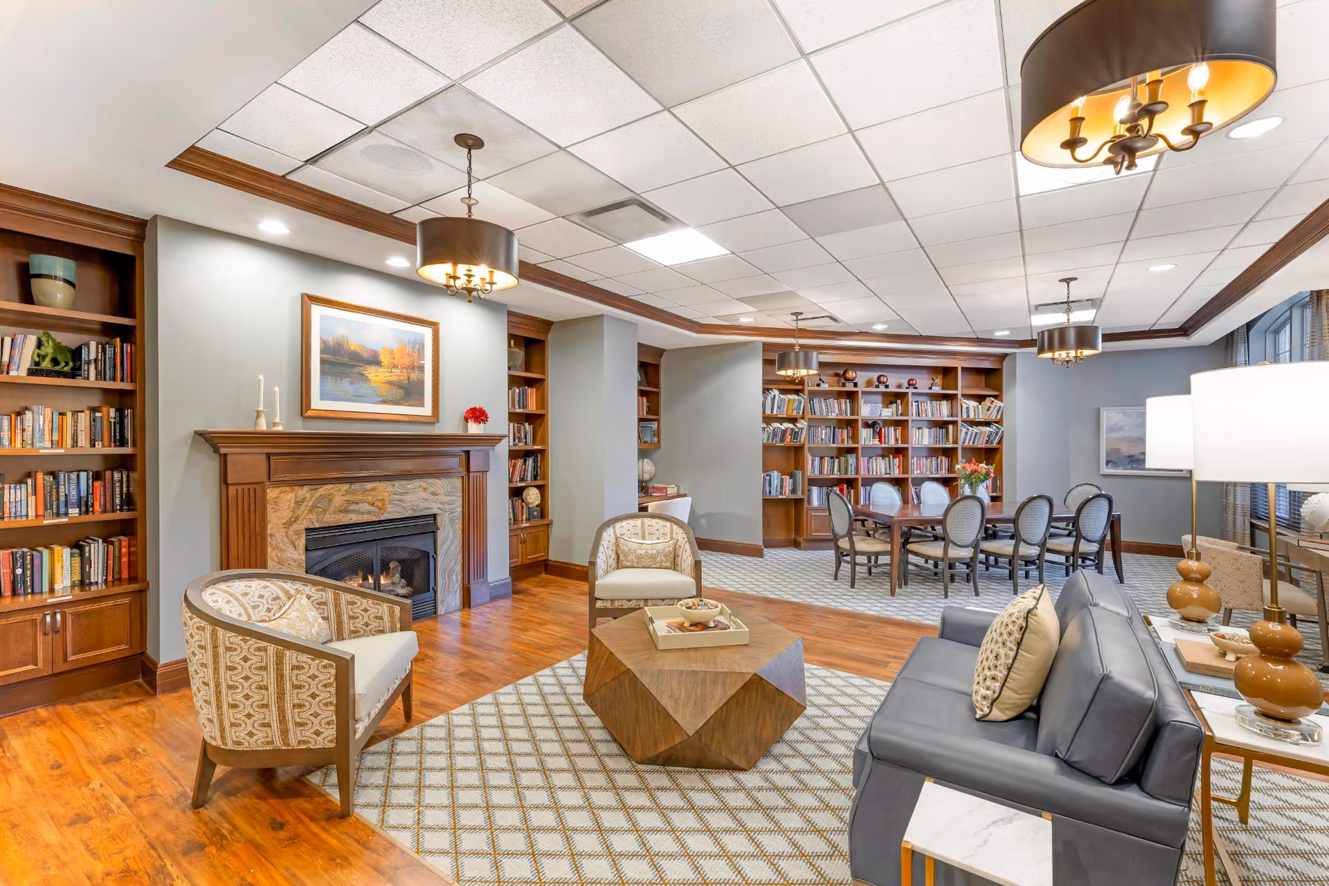 A cozy and well-lit living room area in a senior living facility featuring a fireplace with a painting above it, built-in wooden bookshelves filled with books, comfortable armchairs, a modern geometric coffee table, a gray sofa with decorative pillows, and a dining table with chairs in the background. The room has wooden flooring, patterned rugs, and ceiling lights with drum shades.
