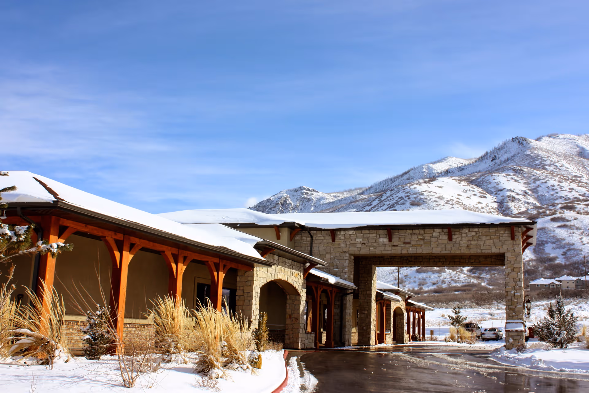 Snow-covered front entrance and porte-cochere of a stone-and-wood building with mountains in the background.