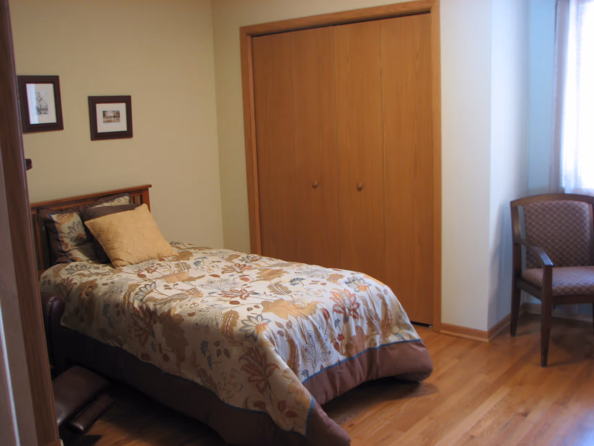 A small bedroom with a single bed covered in a floral patterned comforter and a beige pillow. There is a wooden closet with double doors, a wooden chair with patterned upholstery near a window with sheer curtains, and two framed pictures hanging on the wall above the bed.