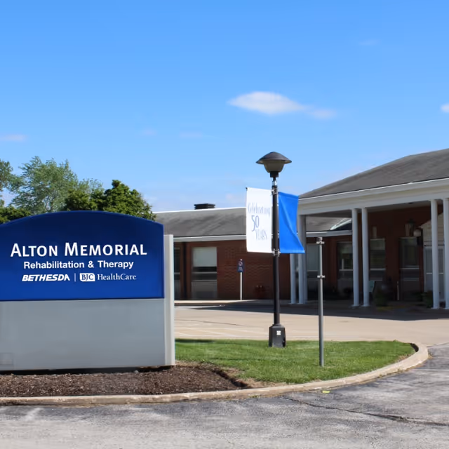 Exterior view of Alton Memorial Rehabilitation & Therapy building with a blue sign displaying the facility name and logos for Bethesda and BJC HealthCare. The building is a single-story brick structure with a covered entrance and a lamppost with banners celebrating 50 years.