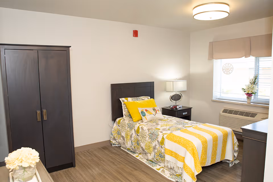 A bright single-occupancy bedroom with a twin bed, floral bedding and yellow accents, a nightstand, wardrobe, and a window with an air conditioner.