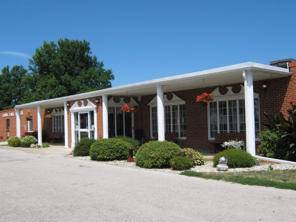 Exterior view of Lenox Care Center, a single-story brick building with white columns supporting a covered walkway. There are hanging flower baskets and well-maintained bushes along the front of the building under a clear blue sky.