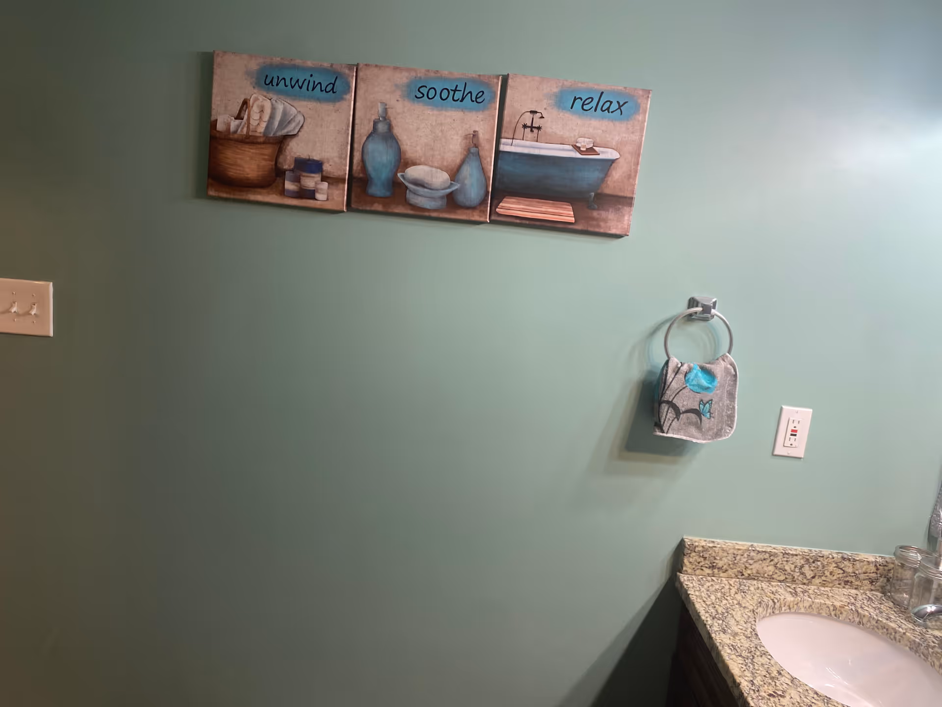 A bathroom wall painted in light green with three decorative pictures hanging, each with words 'unwind', 'soothe', and 'relax' and images of bath items. Below the pictures is a towel ring holding a small towel with a blue flower design. To the right is a granite countertop with a white sink and a glass container.