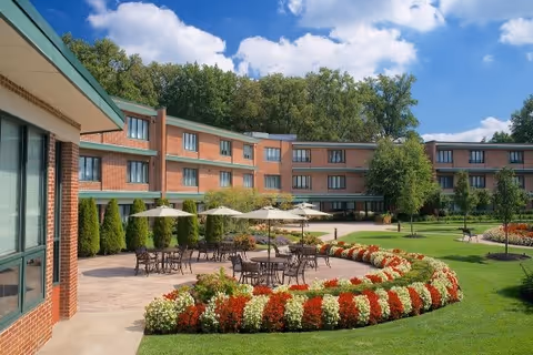 Outdoor patio area of a senior living facility with tables, chairs, and umbrellas surrounded by well-maintained flower beds and green lawns, with a three-story brick building and trees in the background under a partly cloudy sky.