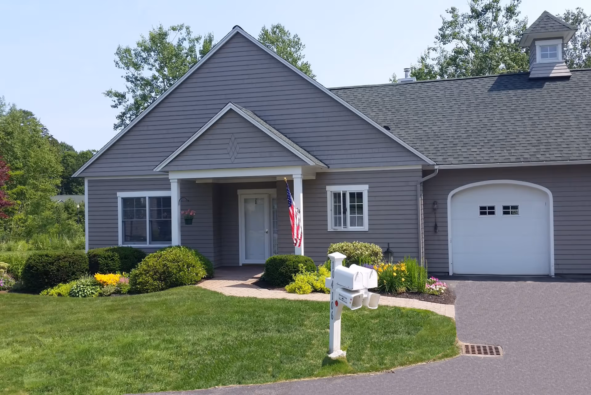 Front exterior of a gray single-story house with a garage, American flag, mailbox, and landscaped lawn.