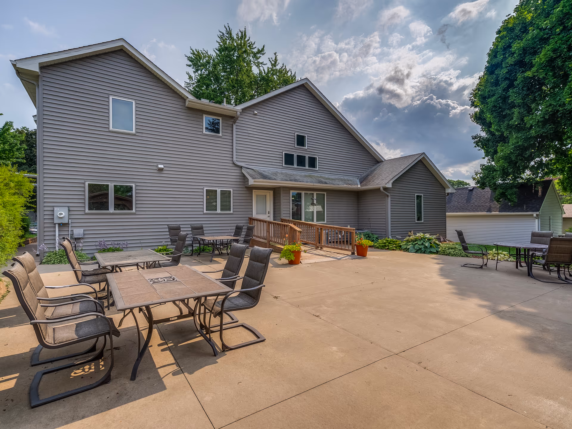 Large concrete patio with tables and chairs in front of a two-story gray-sided house with a small ramp, potted plants and surrounding trees.