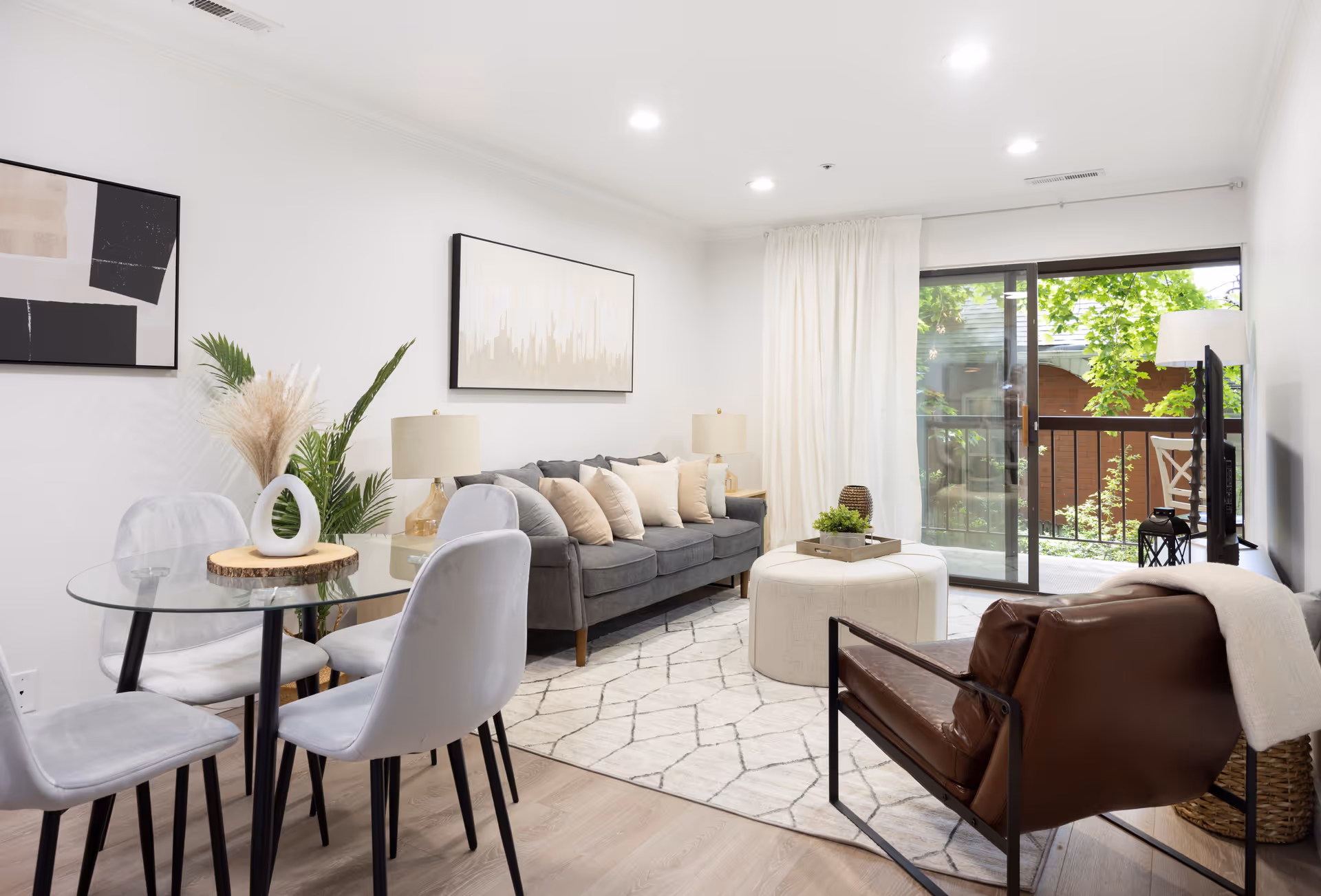A bright and modern living room with a gray sofa adorned with beige and white pillows, a round white ottoman, a brown leather armchair with a white throw, and a glass dining table with four gray chairs. The room features light wood flooring, abstract wall art, a large window with white curtains, and a sliding glass door leading to a balcony with greenery outside.