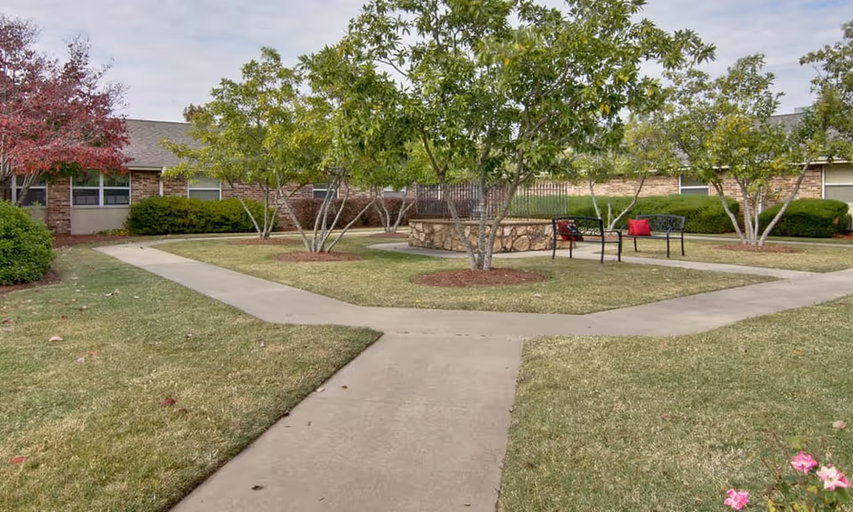 Outdoor courtyard area with concrete walkways intersecting through a grassy lawn, surrounded by small trees and shrubs. There are benches with red cushions near a stone circular structure in the center. The background shows a single-story brick building with windows.