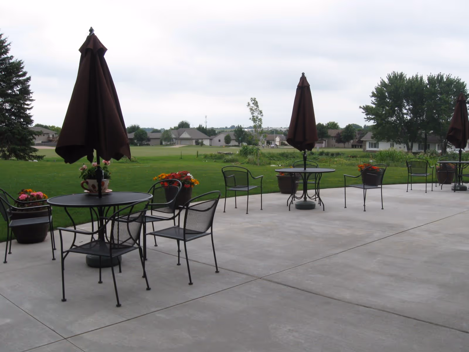 Outdoor patio area with several round metal tables and chairs, each table having a closed dark brown umbrella in the center. There are flower pots with colorful flowers placed near the tables. The patio overlooks a green lawn with trees and houses in the background under a cloudy sky.