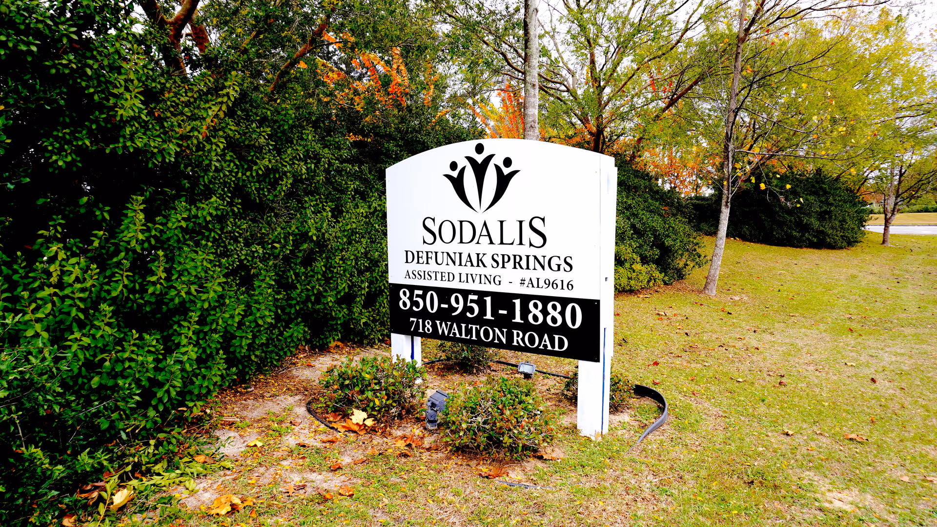 White Sodalis Defuniak Springs assisted living sign on a grassy lawn with trees and shrubs behind it.