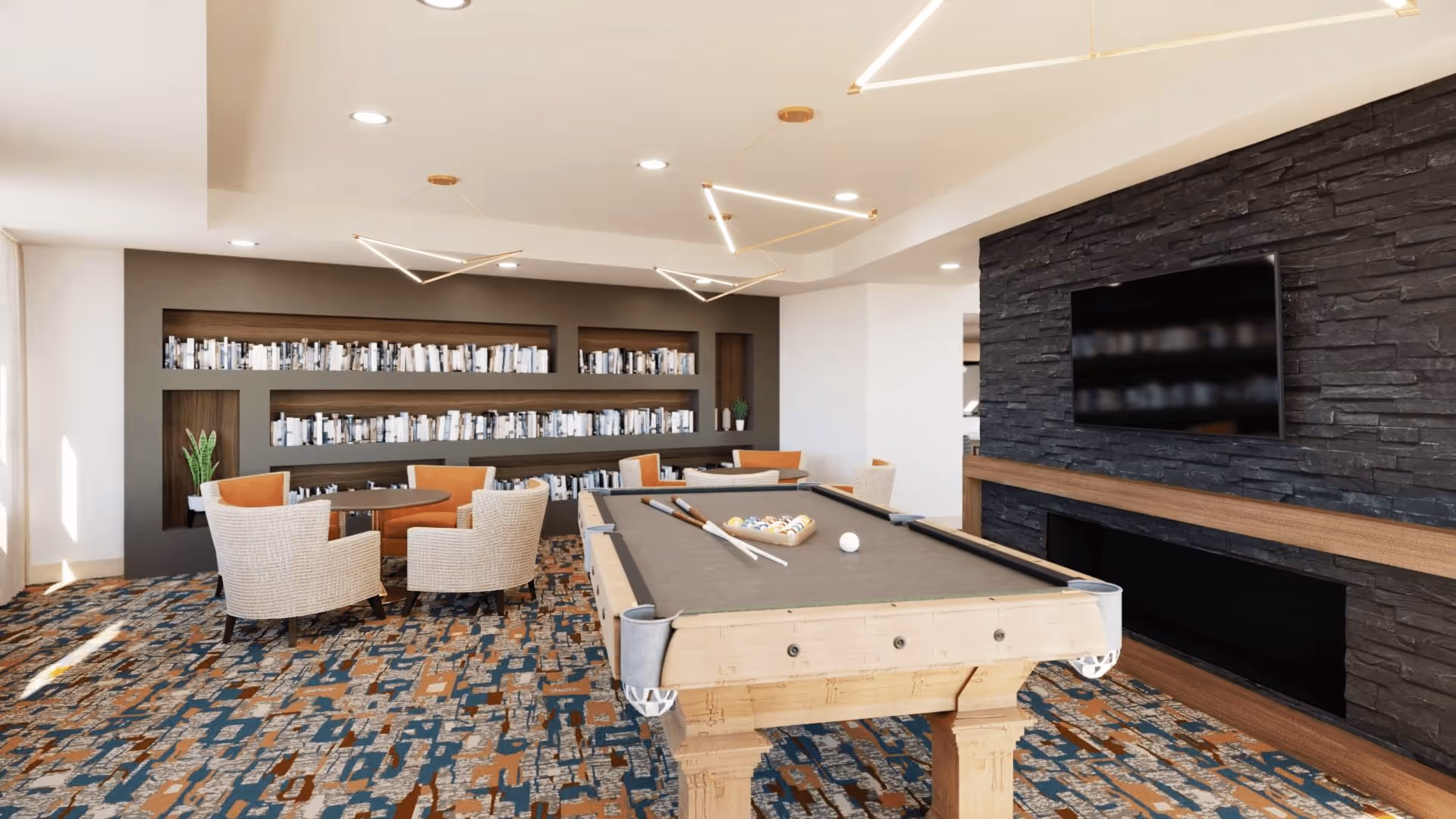 Bright common room with a pool table in the foreground, a round seating area and bookshelves along the back wall, and a wall-mounted TV over a stone fireplace.