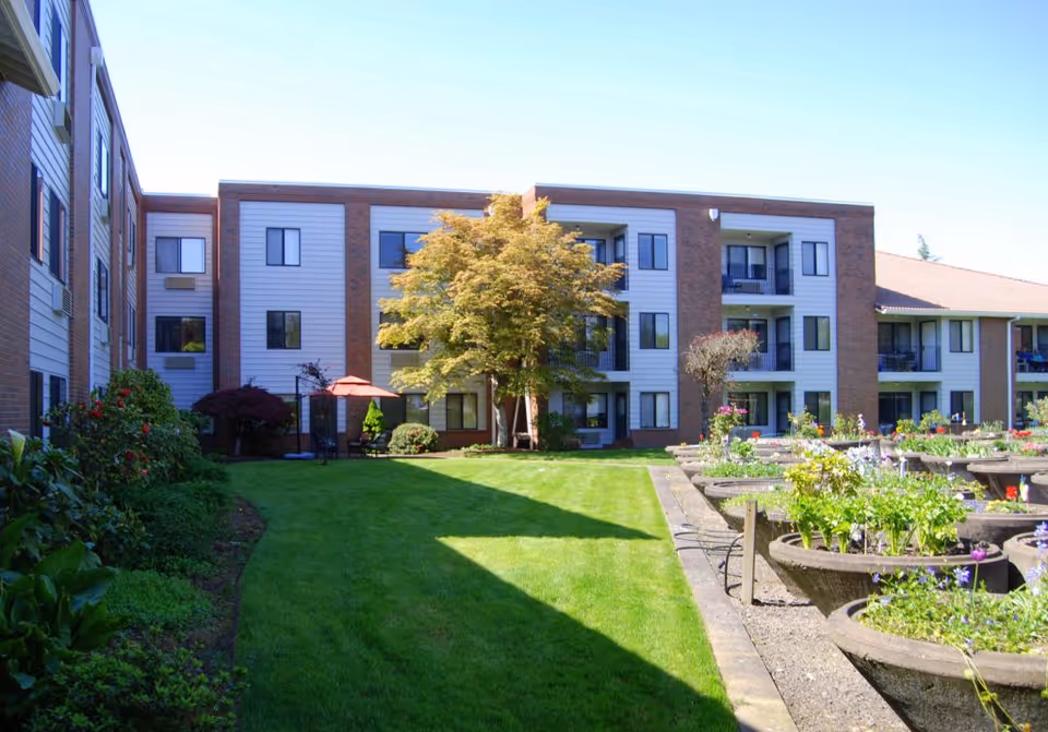 Courtyard with a green lawn, raised flower planters and a three-story senior living building with balconies and windows under a clear blue sky.
