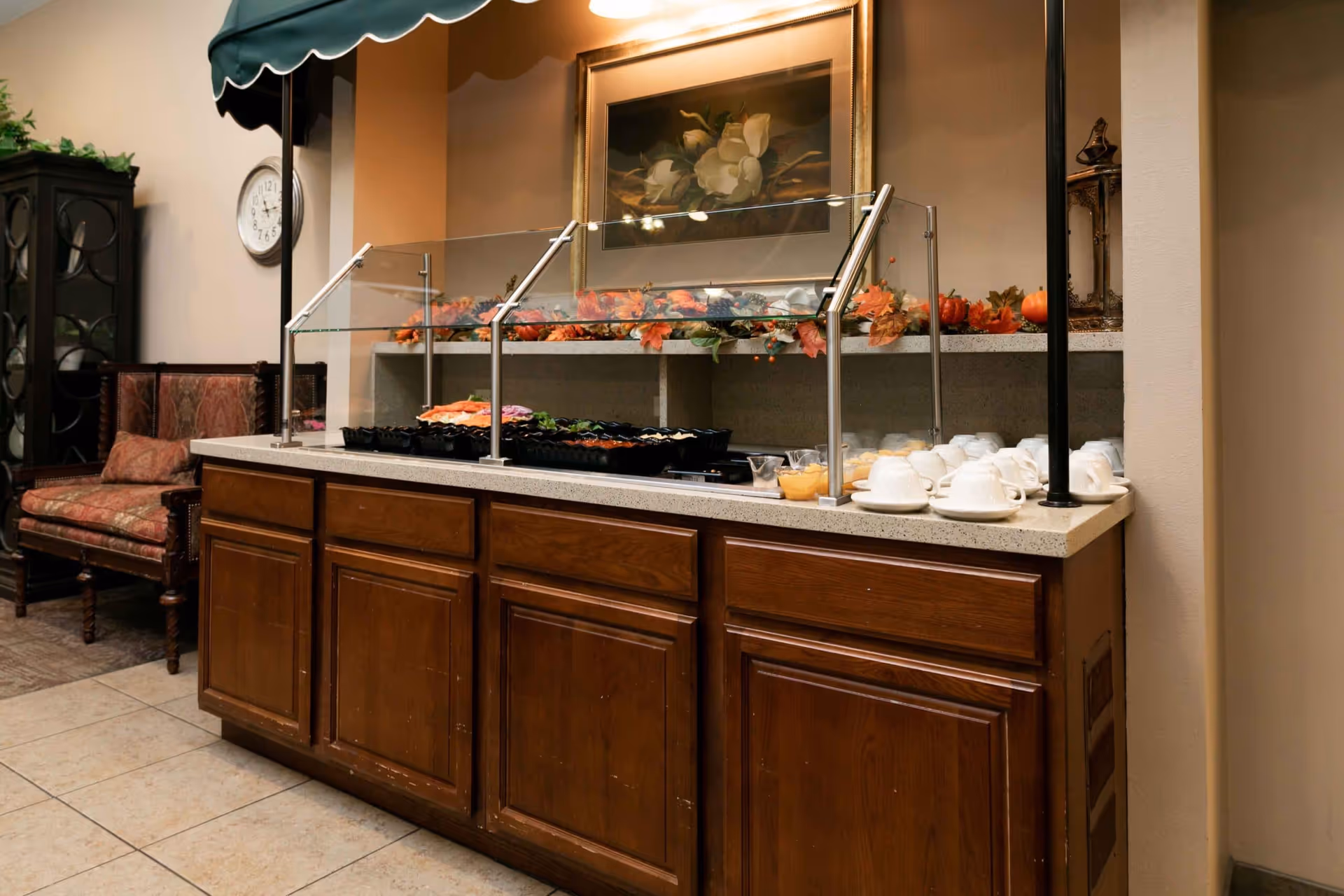 Buffet counter with a glass sneeze guard displaying trays of food, cups and saucers on the side, autumn-themed decorations, a framed floral painting on the wall, and a seating area with upholstered chairs in the background.