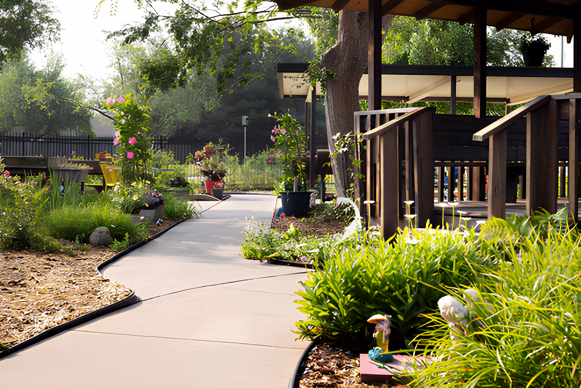 A winding concrete pathway through a garden with various green plants, flowers, and decorative garden ornaments. On the right side, there is a wooden gazebo with railings and a canopy roof. Trees and a black fence are visible in the background under a bright sky.