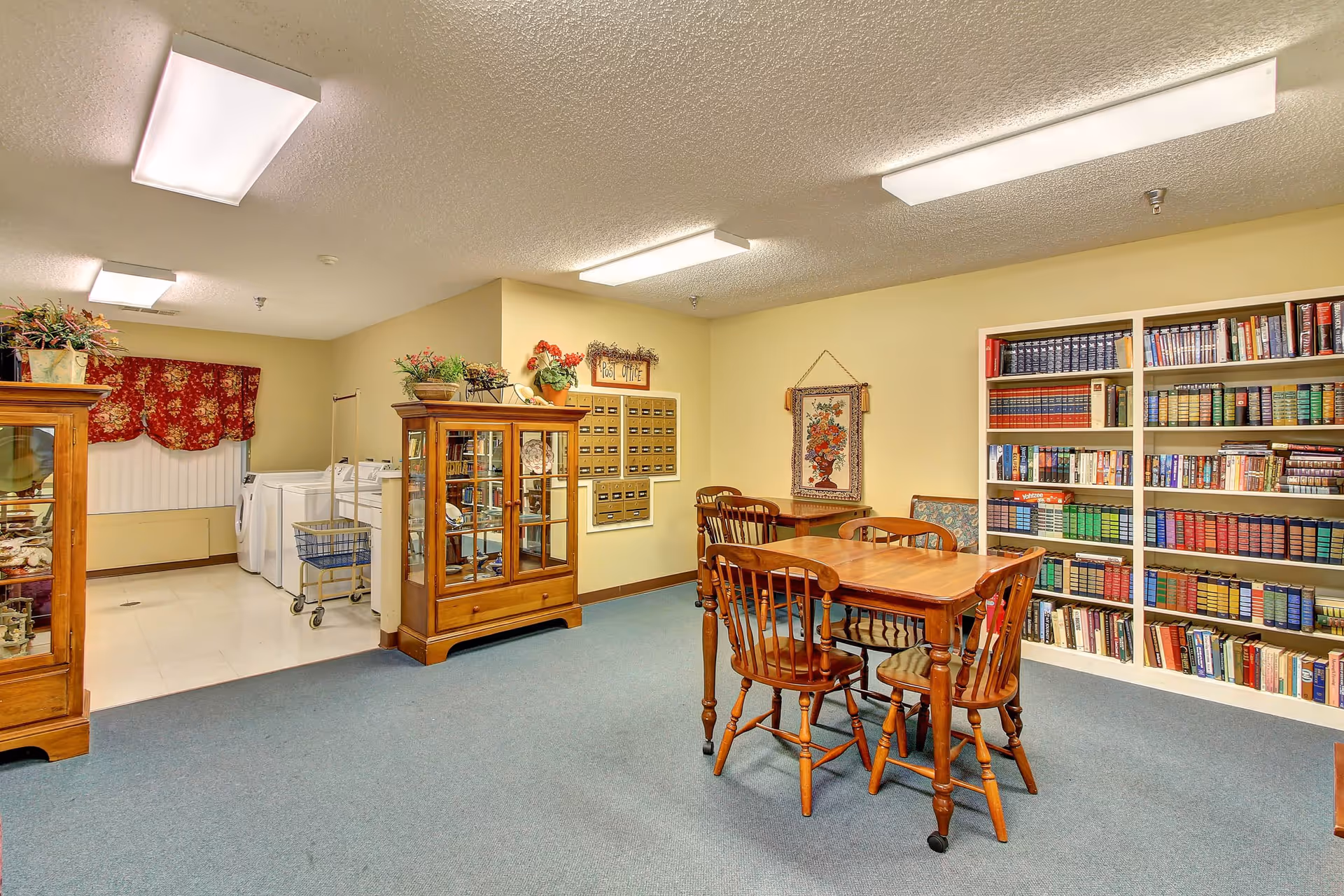 A cozy room with a wooden table and four chairs on a blue carpet. There are bookshelves filled with books on the right wall and a wooden display cabinet with decorative items. In the background, there is a laundry area with a washing machine and dryer, and a wall-mounted set of mailboxes labeled 'Post Office'. The walls are painted light yellow and the ceiling has fluorescent lights.