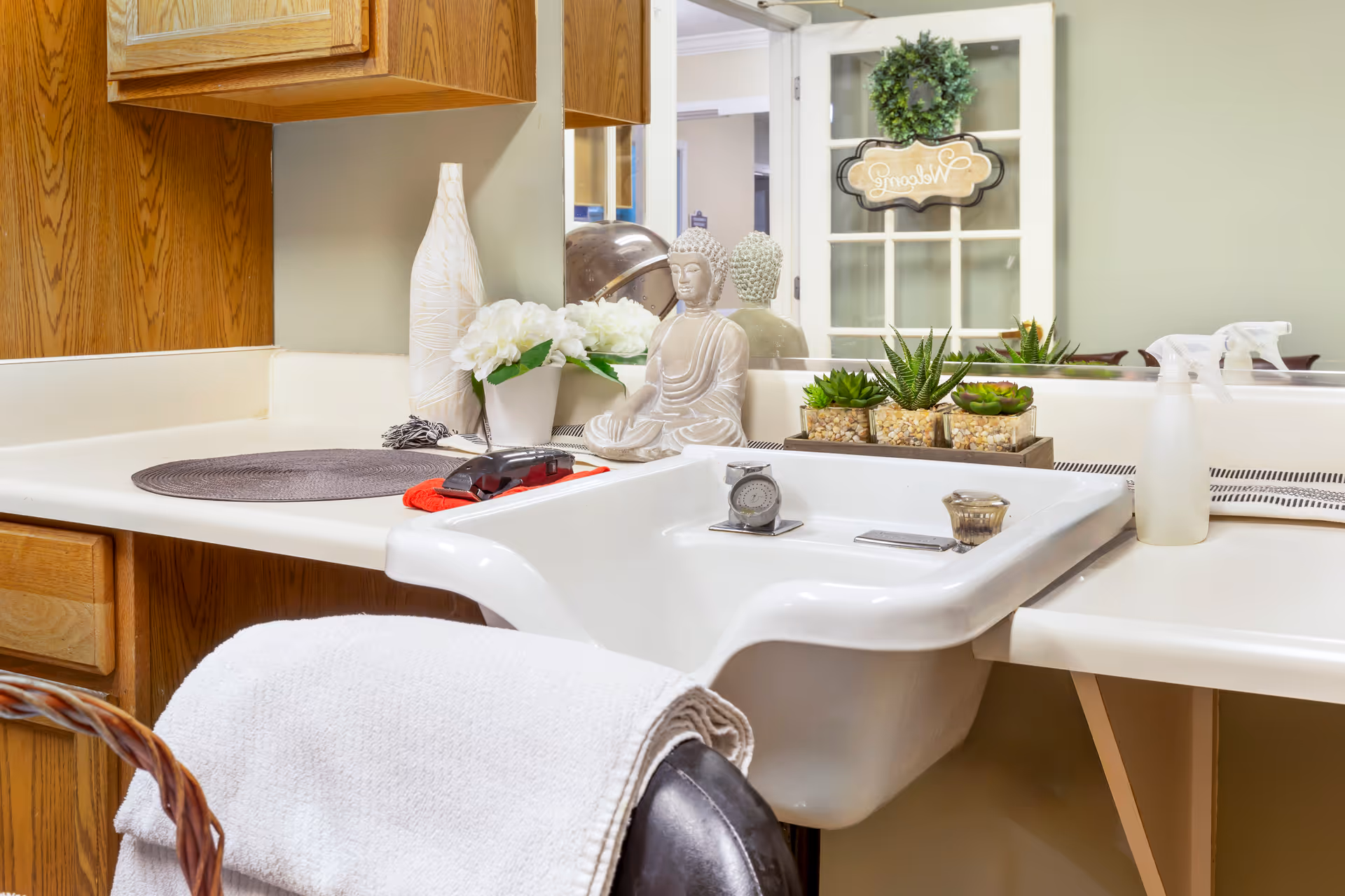 A clean sink and countertop in a senior living facility grooming area decorated with plants, a Buddha statue, and a towel-draped chair.