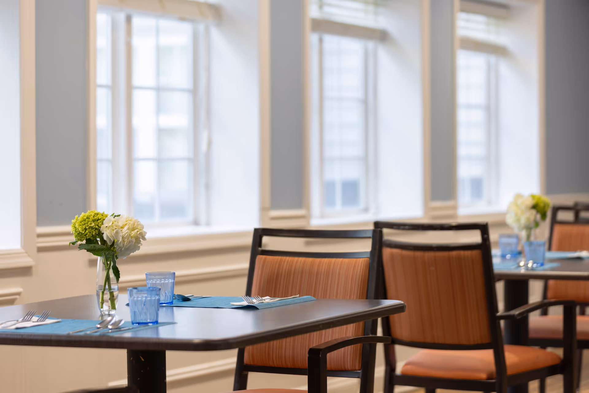A dining area with tables set with blue placemats, blue drinking glasses, silverware, and small vases holding white and green flowers. The room has large windows allowing natural light to fill the space, and chairs with orange cushions and black frames surround the tables.