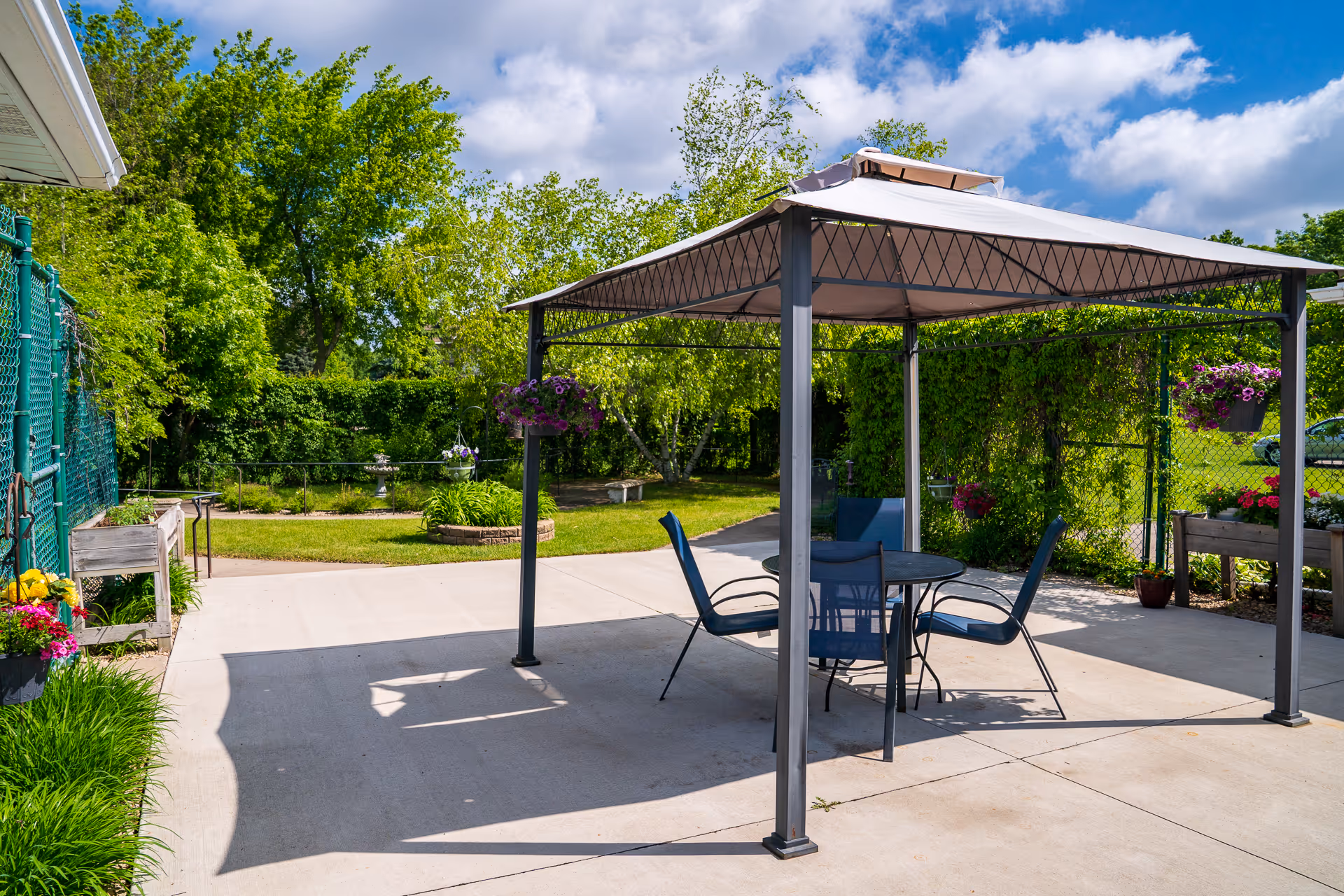 Outdoor patio area with a metal gazebo covering a round glass table and four blue chairs. The patio is surrounded by green trees, bushes, and colorful hanging flower baskets. The sky is partly cloudy with blue patches visible.