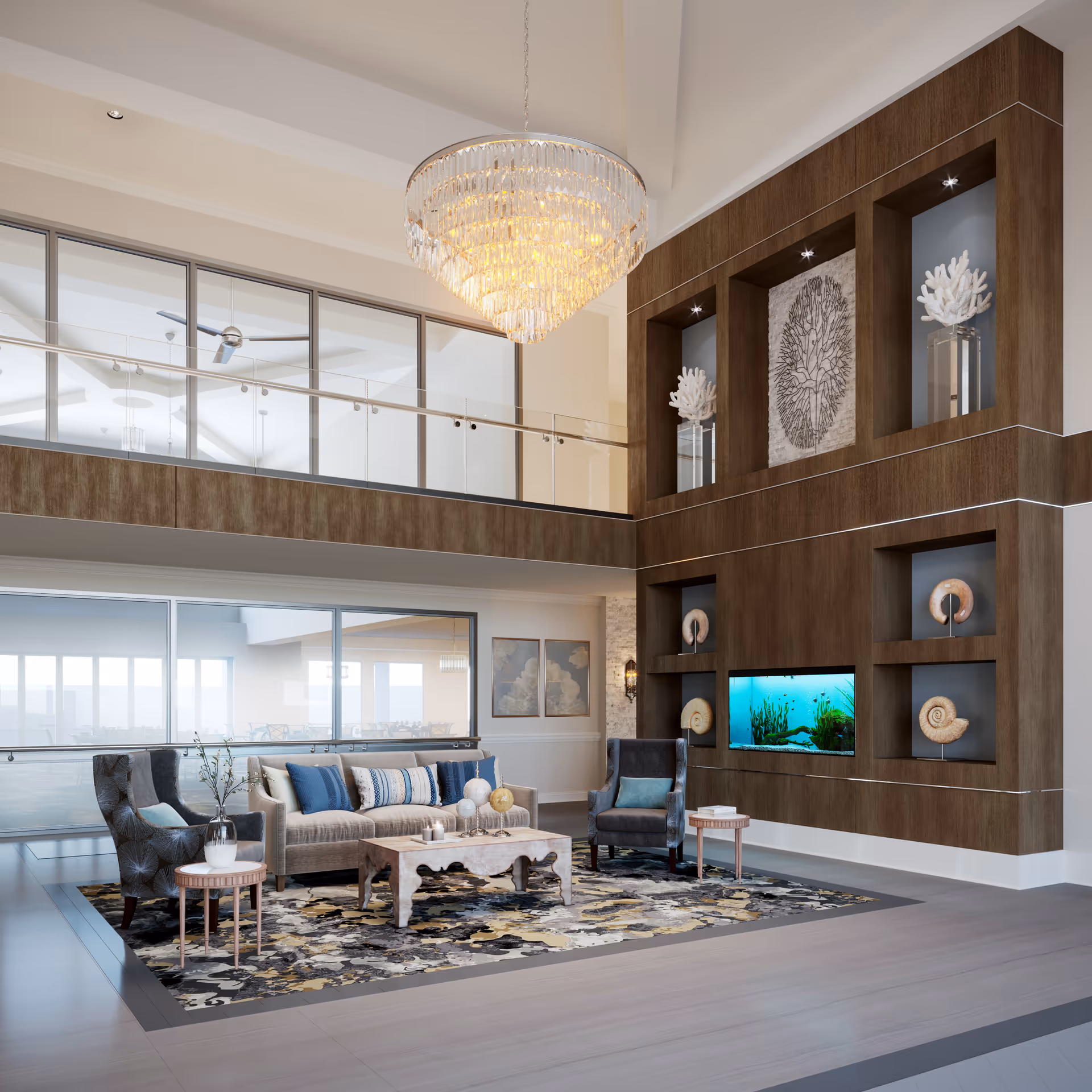 A spacious and elegant living room area with a high ceiling and a large crystal chandelier. The room features a beige sofa with blue and patterned cushions, two dark upholstered armchairs, a wooden coffee table, and two small side tables. The back wall has built-in wooden shelves with decorative coral sculptures and a built-in aquarium. Large windows and glass railings overlook an adjacent room with tables and chairs.