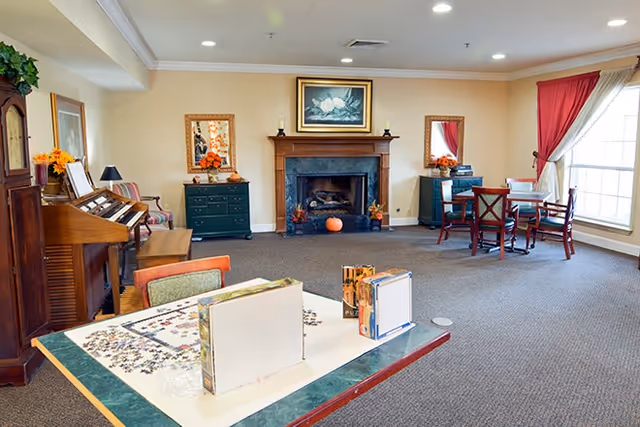 Bright communal lounge with a fireplace, tables and chairs, a piano, and a puzzle table near a large window.