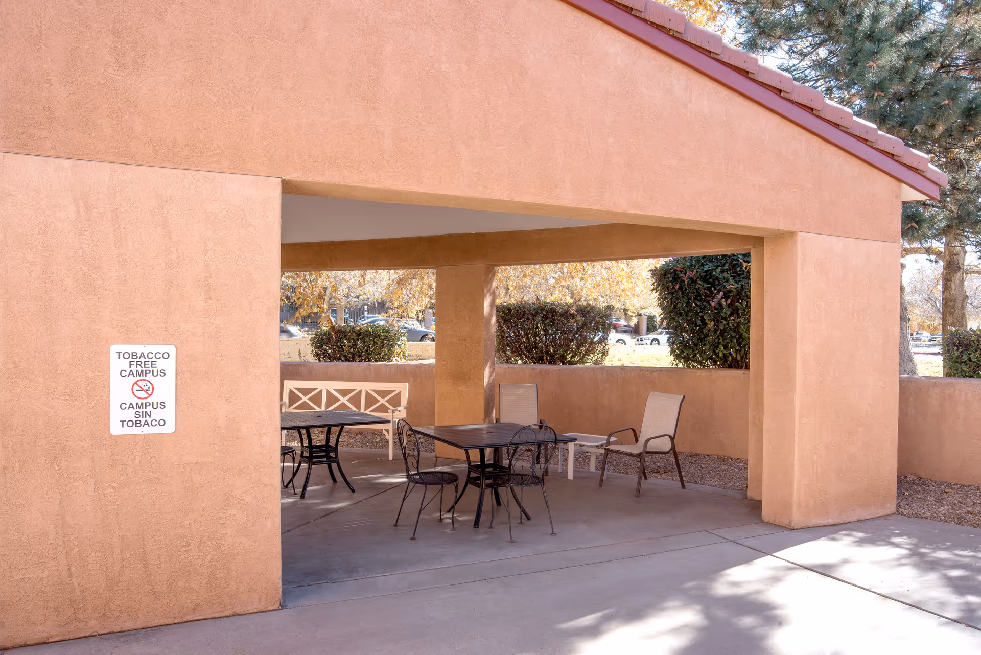 Outdoor covered seating area with several chairs and tables under a tan stucco structure. There are bushes and trees in the background, and a sign on the wall reads 'TOBACCO FREE CAMPUS CAMPUS SIN TABACO'.