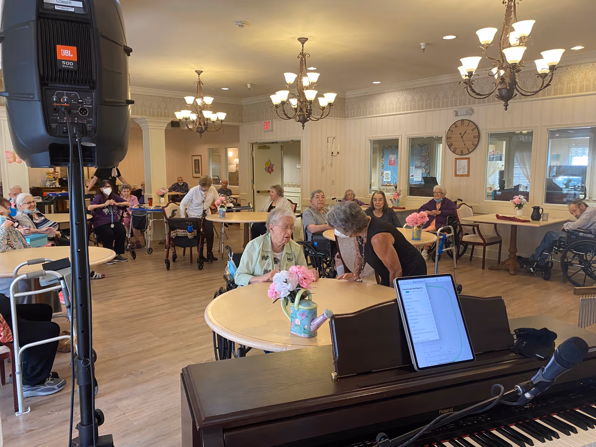 A group of elderly people seated around tables in a well-lit room with chandeliers. Some are in wheelchairs, and a woman is leaning over to speak with an elderly woman at the front table. A piano with a microphone and a tablet is visible in the foreground, and a large speaker is on the left side of the image. The room has light-colored walls and wooden floors, with pink flower arrangements on the tables.