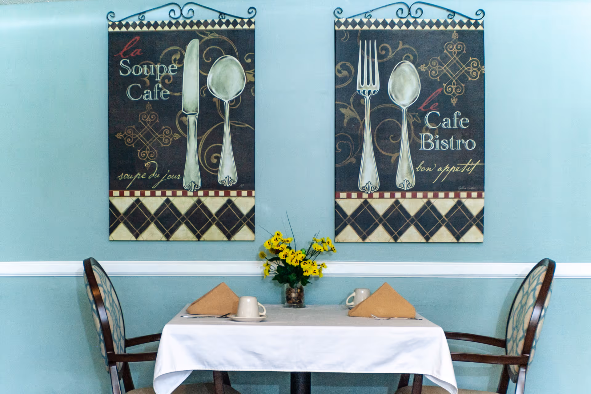 A small dining table set for two with a white tablecloth, two beige napkins folded in triangles, two coffee cups on saucers, and silverware. A vase with yellow flowers is centered on the table. Behind the table, two decorative wall hangings feature large images of utensils and French text, mounted on a light blue wall with white trim.