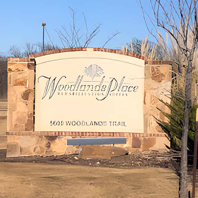 Stone monument entrance sign reading 'Woodlands Place Rehabilitation Suites' with the address carved below set on a grassy landscaped area.