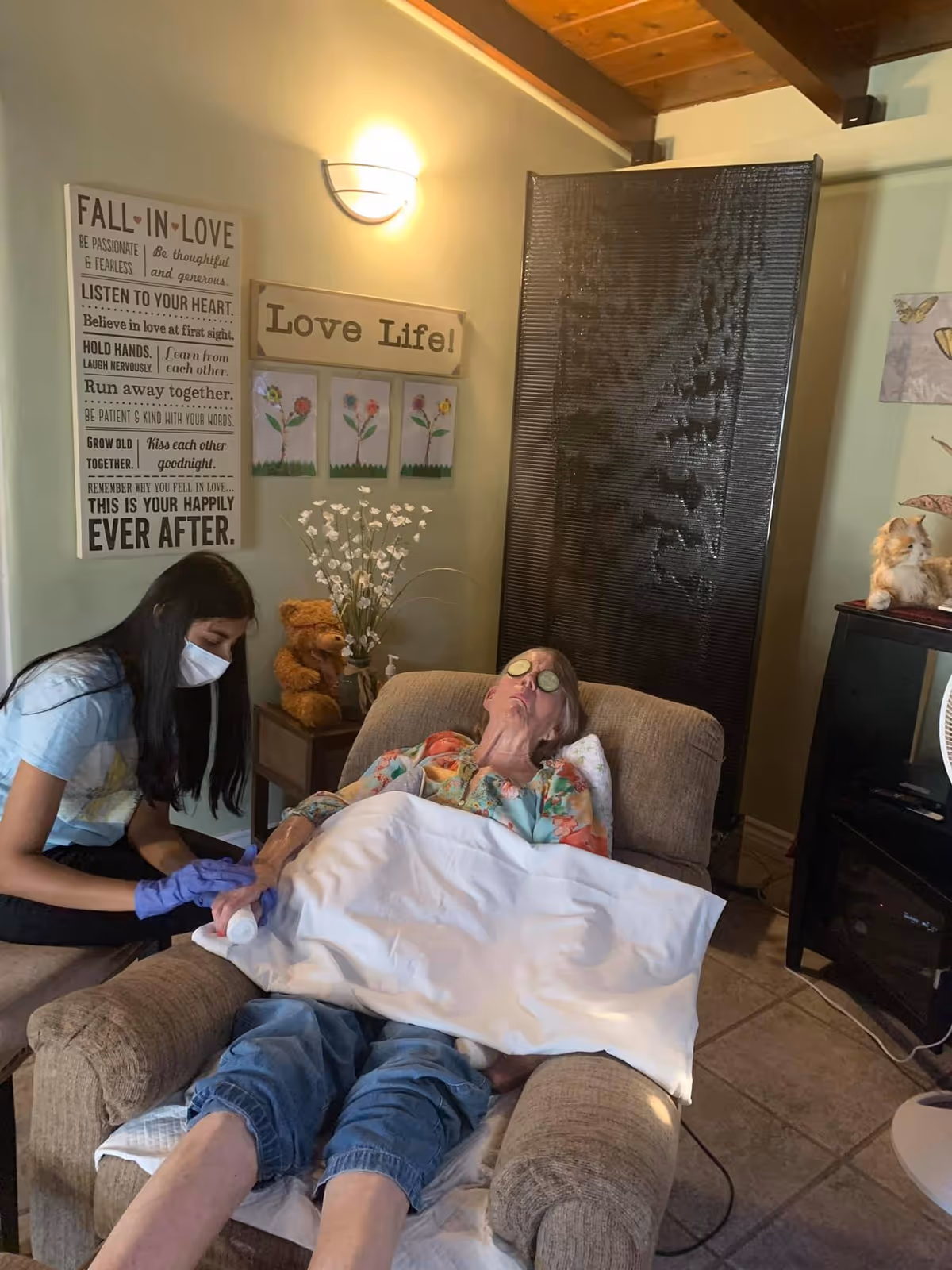 An elderly woman reclines in a cushioned armchair with a white blanket over her legs and cucumber slices on her eyes, receiving care from a young woman wearing a mask and gloves. The room has warm lighting, wall decorations including a sign that says 'Love Life!' and another with inspirational text, a teddy bear on a small table, and a black water feature behind the chair.