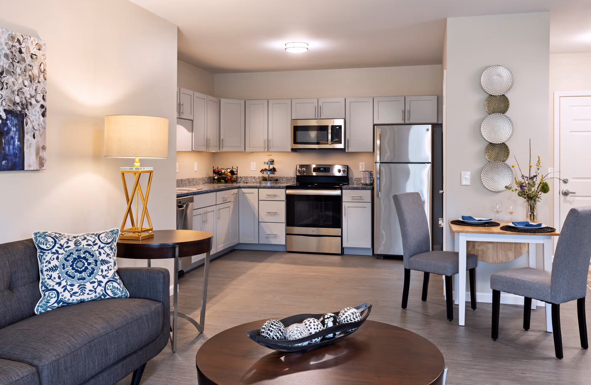 A modern open-concept living space featuring a kitchen with white cabinets, stainless steel appliances including a refrigerator, stove, and microwave. Adjacent to the kitchen is a small dining area with a wooden table set for two with gray upholstered chairs. In the foreground, there is a dark gray sofa with a blue and white patterned pillow, a round wooden coffee table with decorative spheres, and a side table with a lamp. The walls are light-colored with decorative wall art and plates.