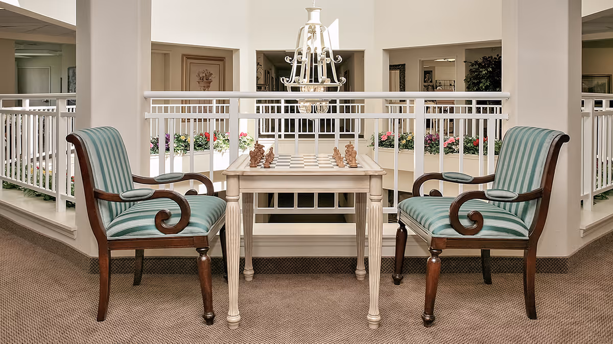 Two striped upholstered armchairs flank a small table with a chessboard in a bright interior common area.