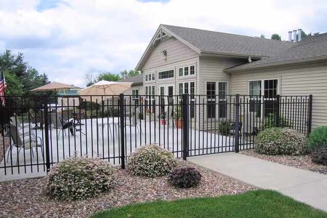 Outdoor patio area at Copperleaf Memory Care of Schofield featuring a fenced-in concrete patio with tables, chairs, and umbrellas. The patio is adjacent to a beige building with multiple windows and a peaked roof. There are landscaped bushes and rocks surrounding the patio area.