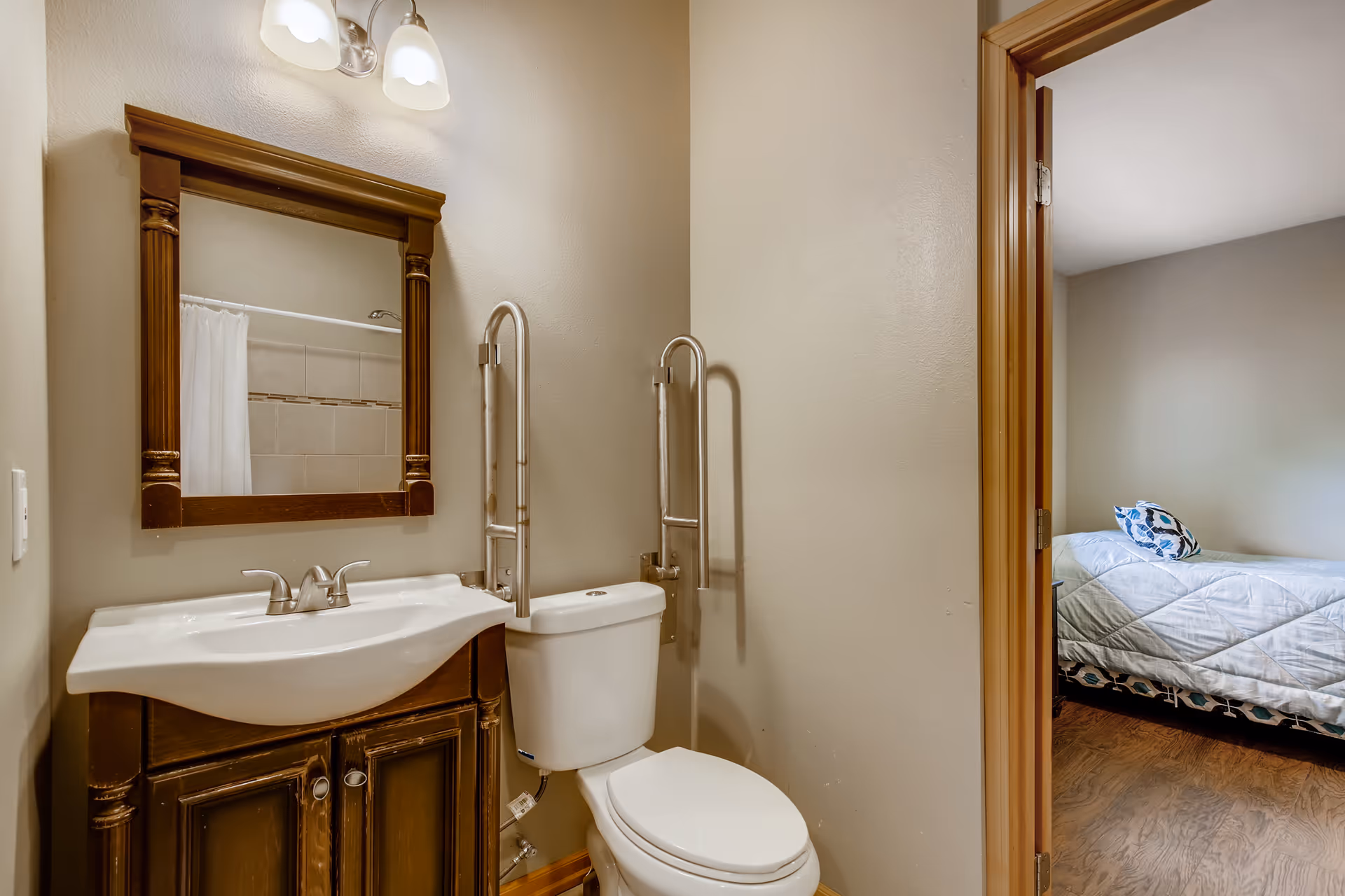 Small bathroom with a sink and wooden-framed mirror, a toilet with stainless grab bars, and a doorway leading to a bedroom.