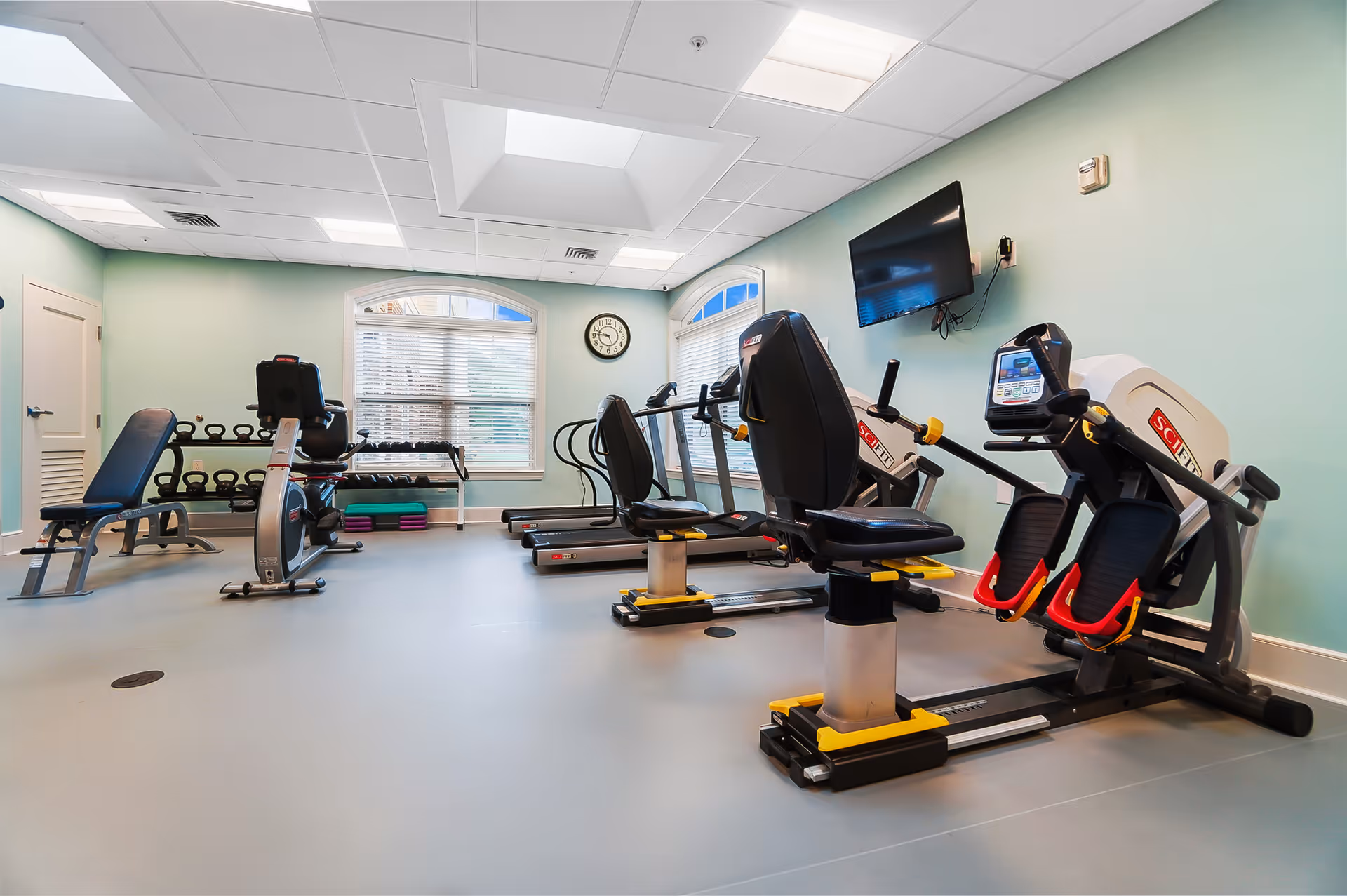 Exercise room with recumbent bikes, treadmills, a weight bench and free weights under a wall-mounted TV.