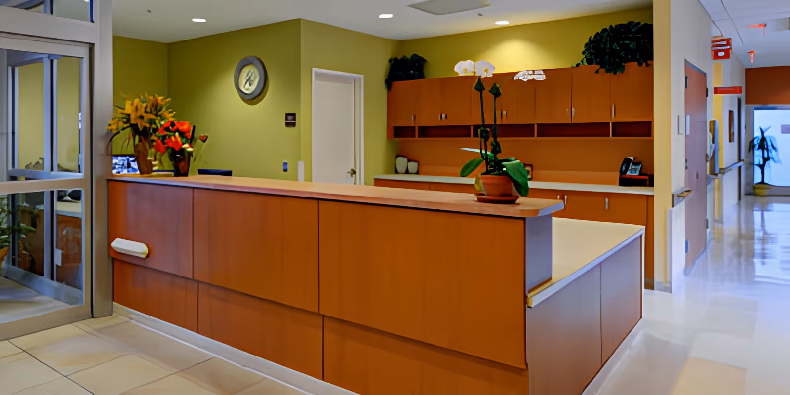Reception desk area in a senior living facility with wooden cabinetry, a potted orchid plant on the counter, a clock on the green wall, and a hallway extending to the right.