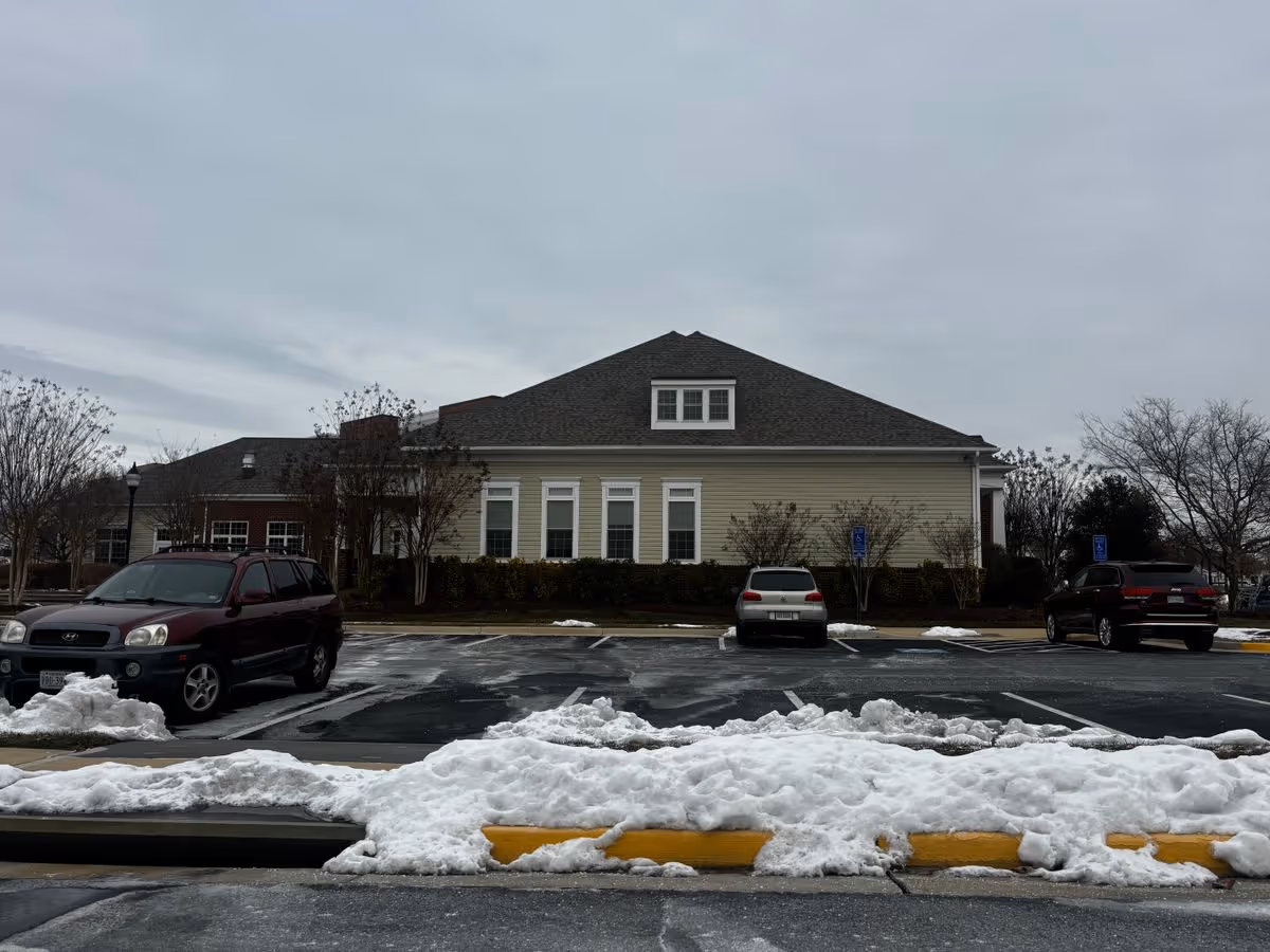 Front view of a single-story light-yellow building with a parking lot, parked cars, and piles of snow along the curb.