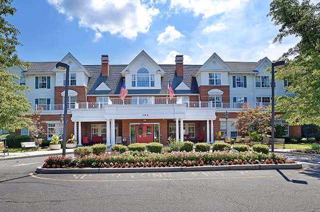 Front view of a three-story red-brick and white senior living building with columns, American flags, landscaped flower beds, and a drive-up entrance.