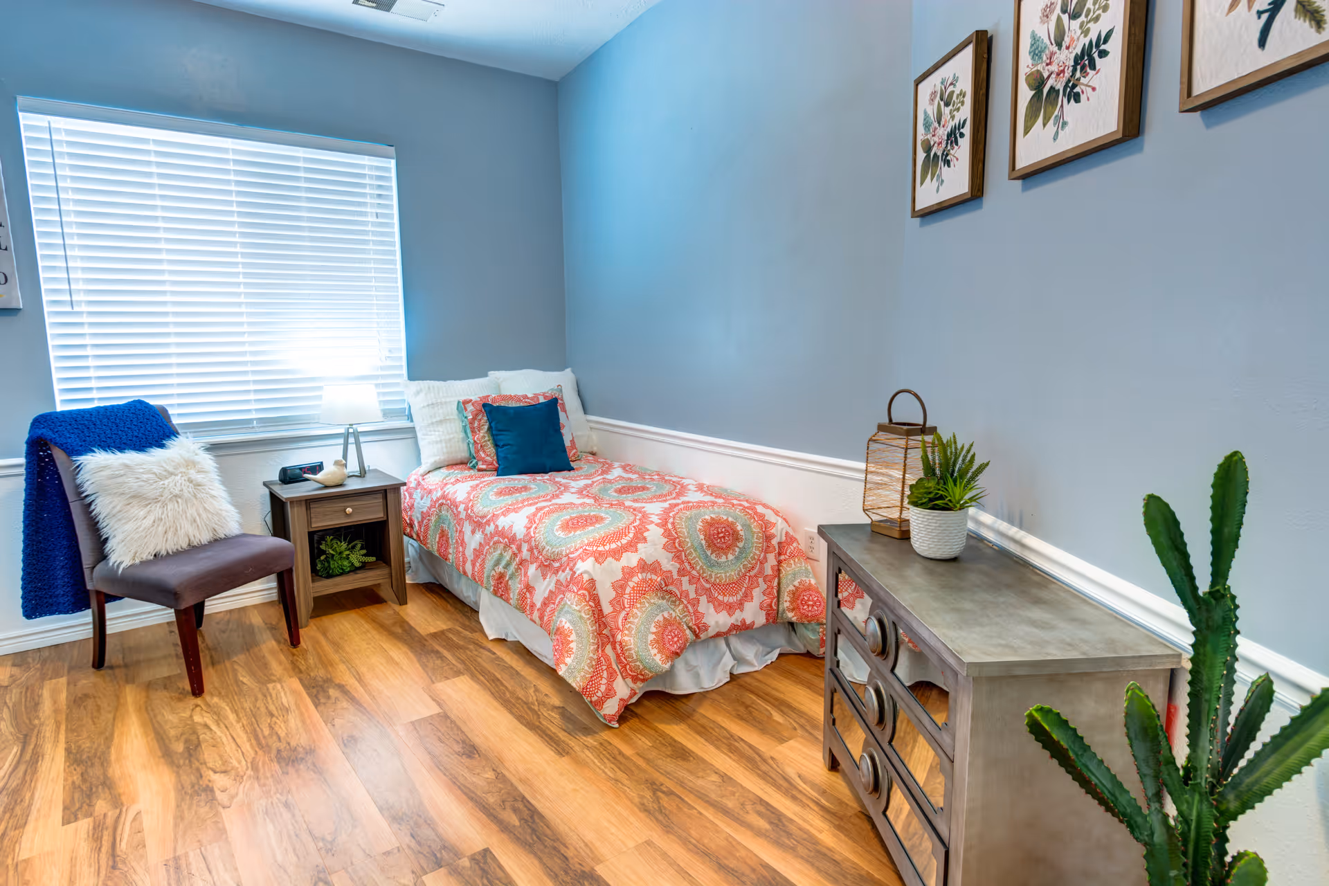 Small furnished bedroom with a single bed, nightstand, chair, dresser, and wall art.