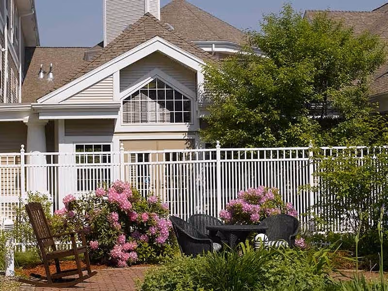 Sunny outdoor courtyard with pink flowering bushes, a table and chairs, and a white fence in front of a building facade.