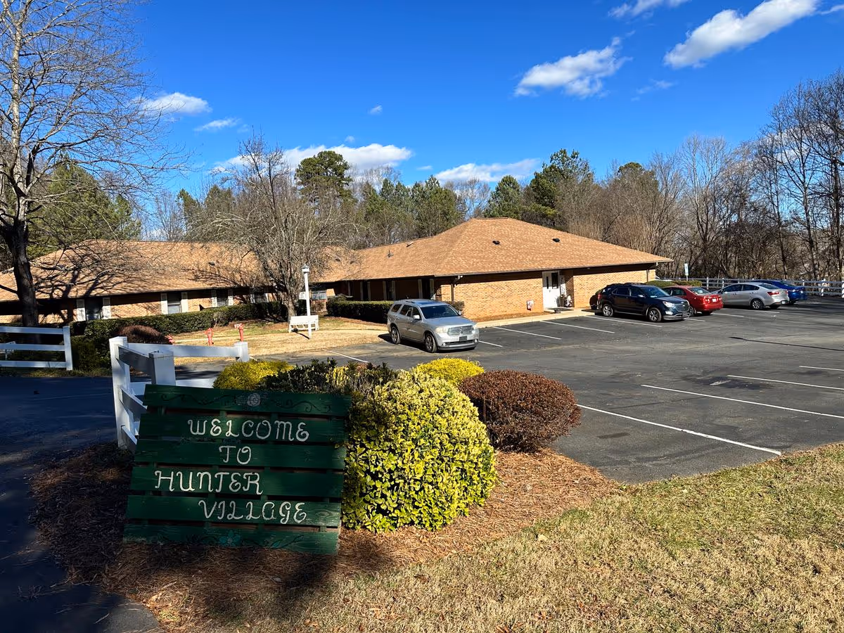 Front exterior of a single-story brick building with a parking lot, cars, landscaping, and a wooden sign reading 'Welcome to Hunter Village'.