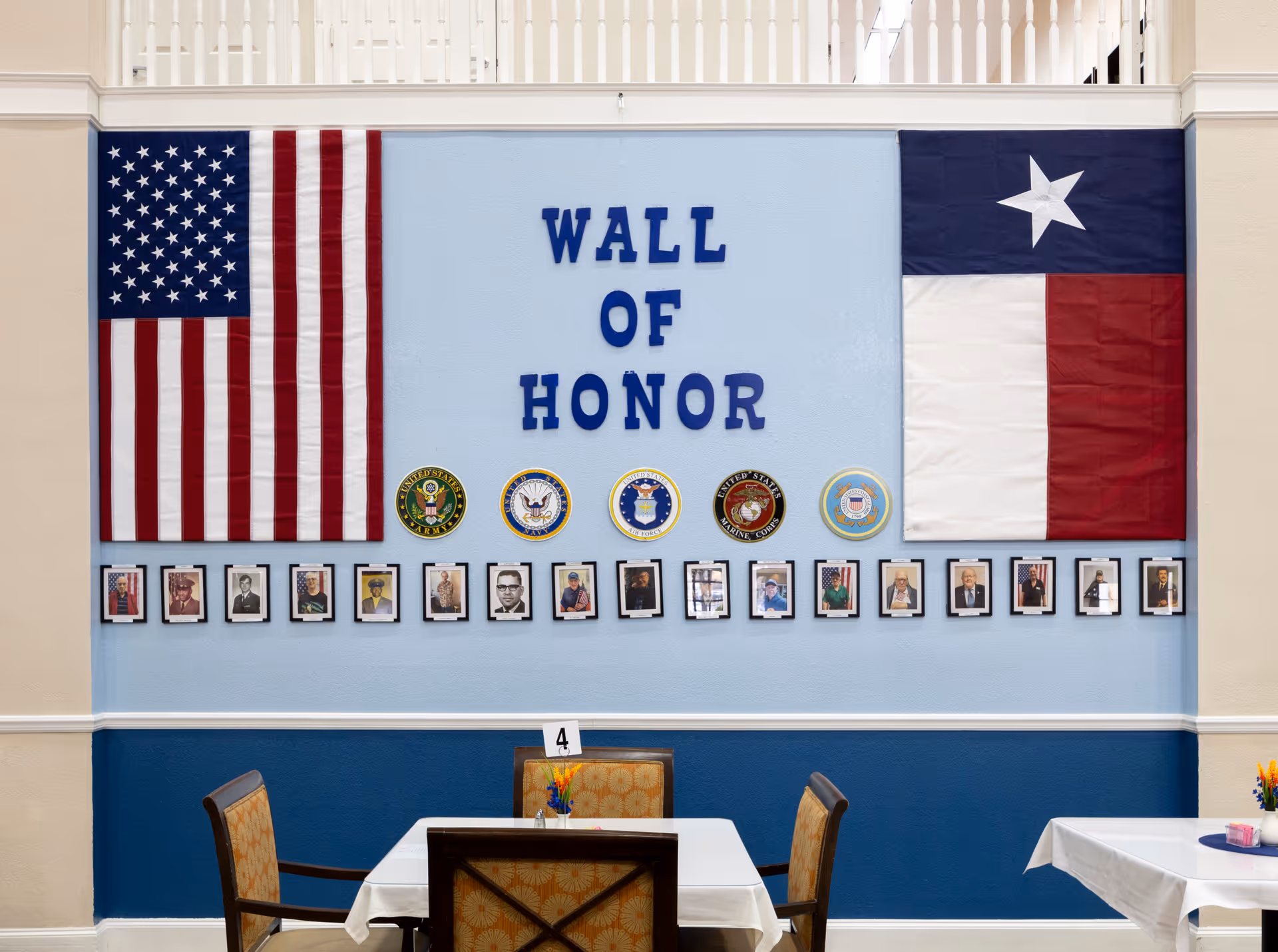 Dining area with a 'Wall of Honor' display showing American and Texas flags, military seals and framed photos above tables and chairs.