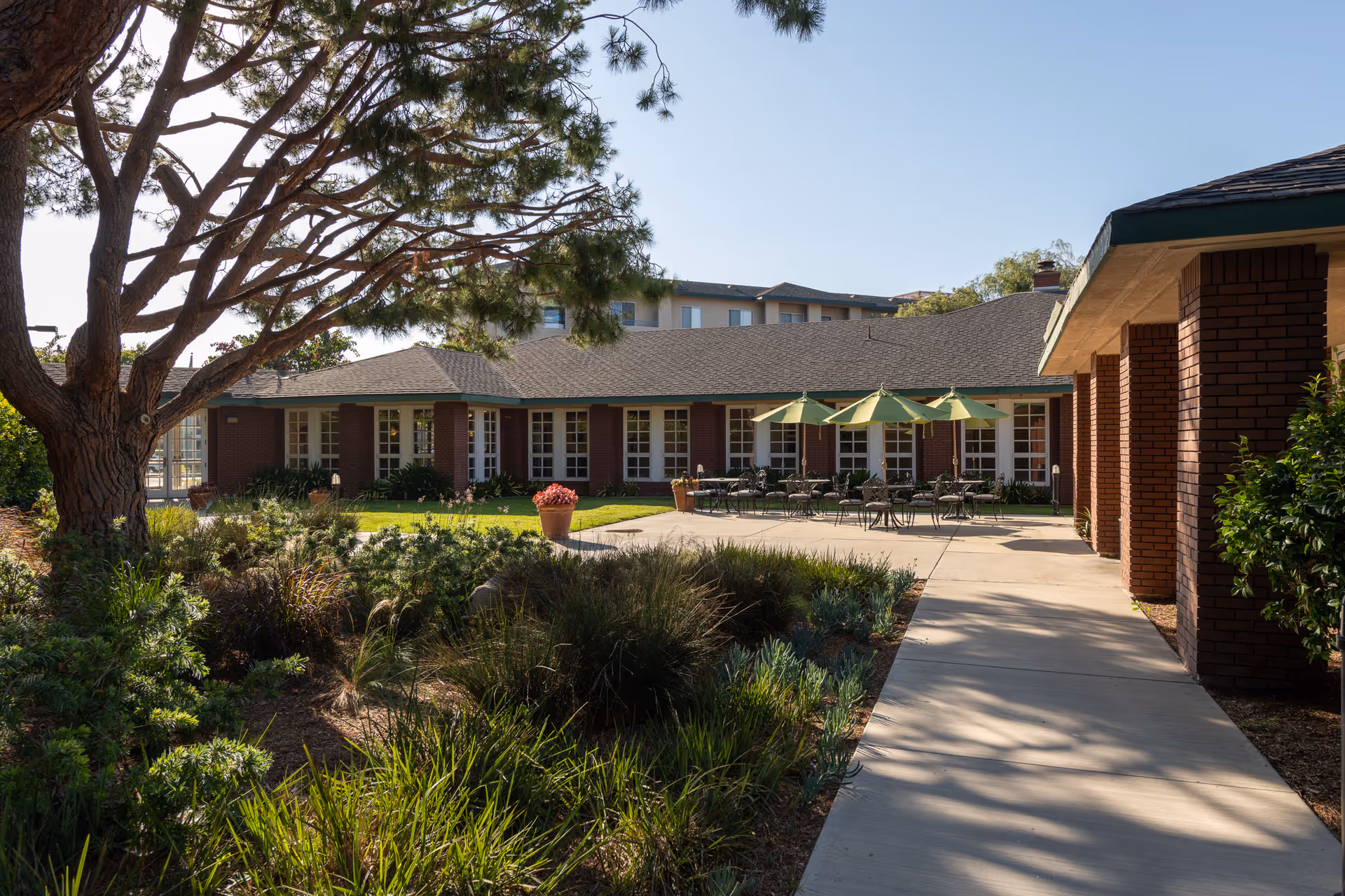 Sunlit landscaped courtyard with patio tables and green umbrellas in front of a low brick retirement community building.