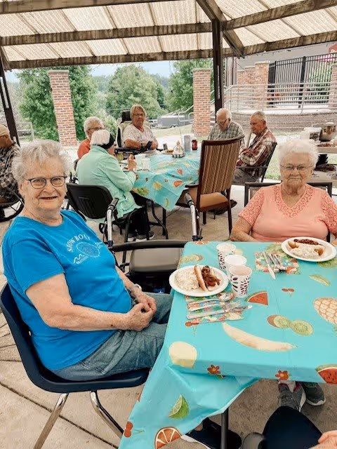 A group of elderly people sitting at tables covered with turquoise tablecloths decorated with fruit patterns, enjoying a meal outdoors under a covered patio. Two women are seated closest to the camera, smiling, with plates of food and cups in front of them. Other seniors are seated at tables in the background with trees and brick pillars visible.