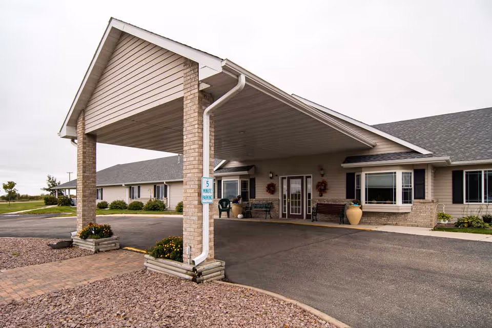 Exterior view of the entrance to a single-story senior living facility with a covered drop-off area supported by brick columns, a paved driveway, and landscaped areas with flowers and shrubs.