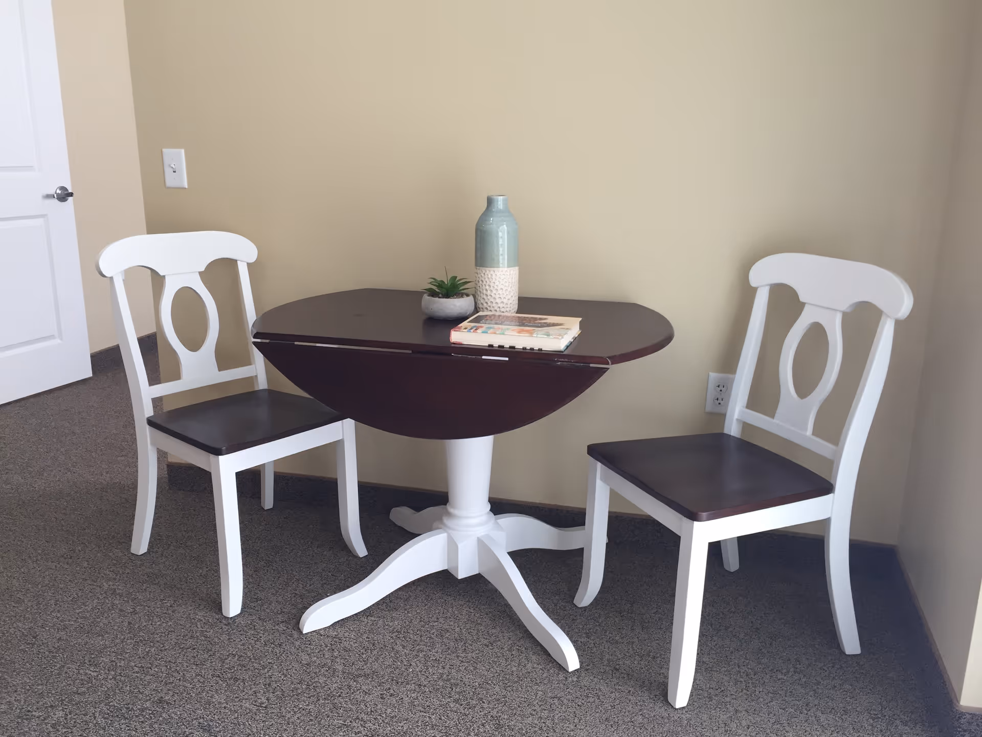 A small dining area with a dark wood oval table with white pedestal base and two matching chairs with white frames and dark wood seats. On the table are a small potted plant, a decorative vase, and a book. The room has beige walls and carpeted floor.