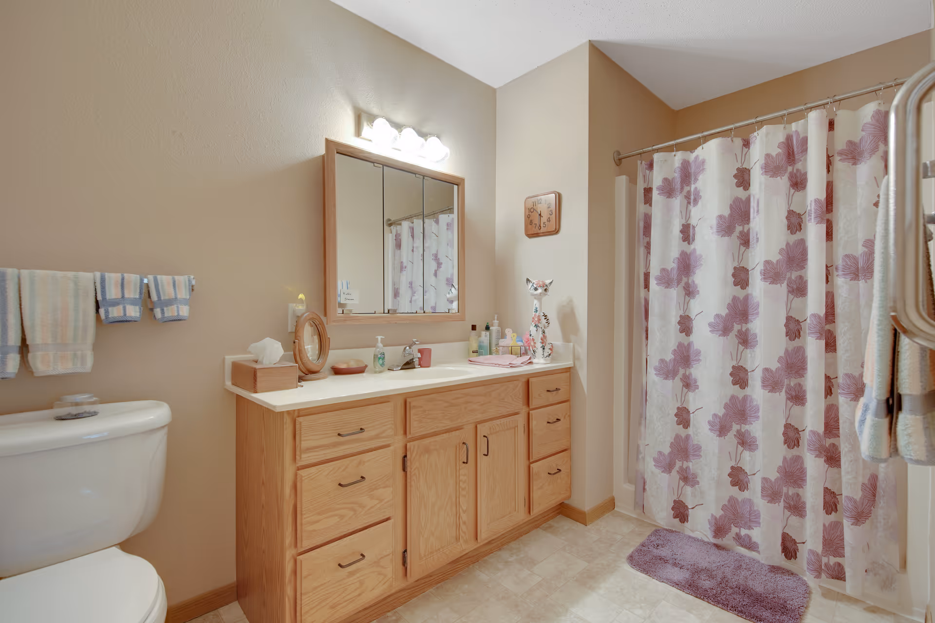Bathroom with a wooden vanity and mirror, toilet, towels, and a shower with a purple floral curtain.