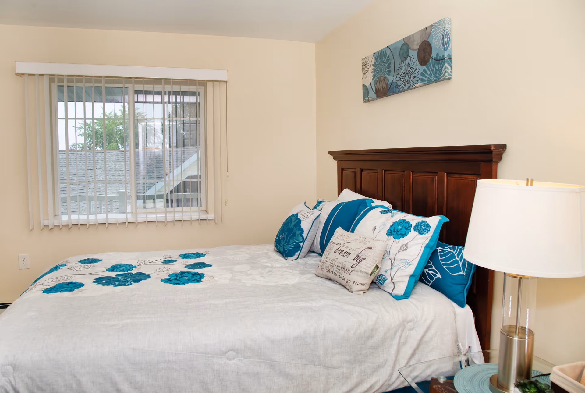 A bedroom with a bed featuring a dark wooden headboard, white and teal floral bedding, and multiple decorative pillows. There is a window with vertical blinds on the left wall and a modern table lamp on a glass nightstand to the right of the bed. A piece of floral artwork hangs above the headboard.